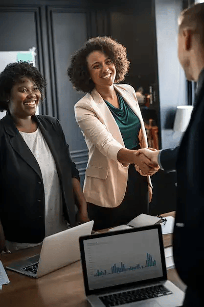 Two forward facing smiling business people where one is shaking hands over table with another business person