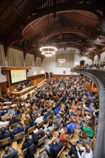 a theatre of people facing a stage with a projector screen behind a panel of speakers