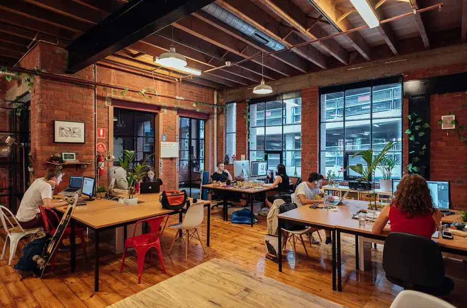 an open, no-cubicle office with people working at multiple tables and two large windows at the far wall letting in natural light for the scattered potted plants